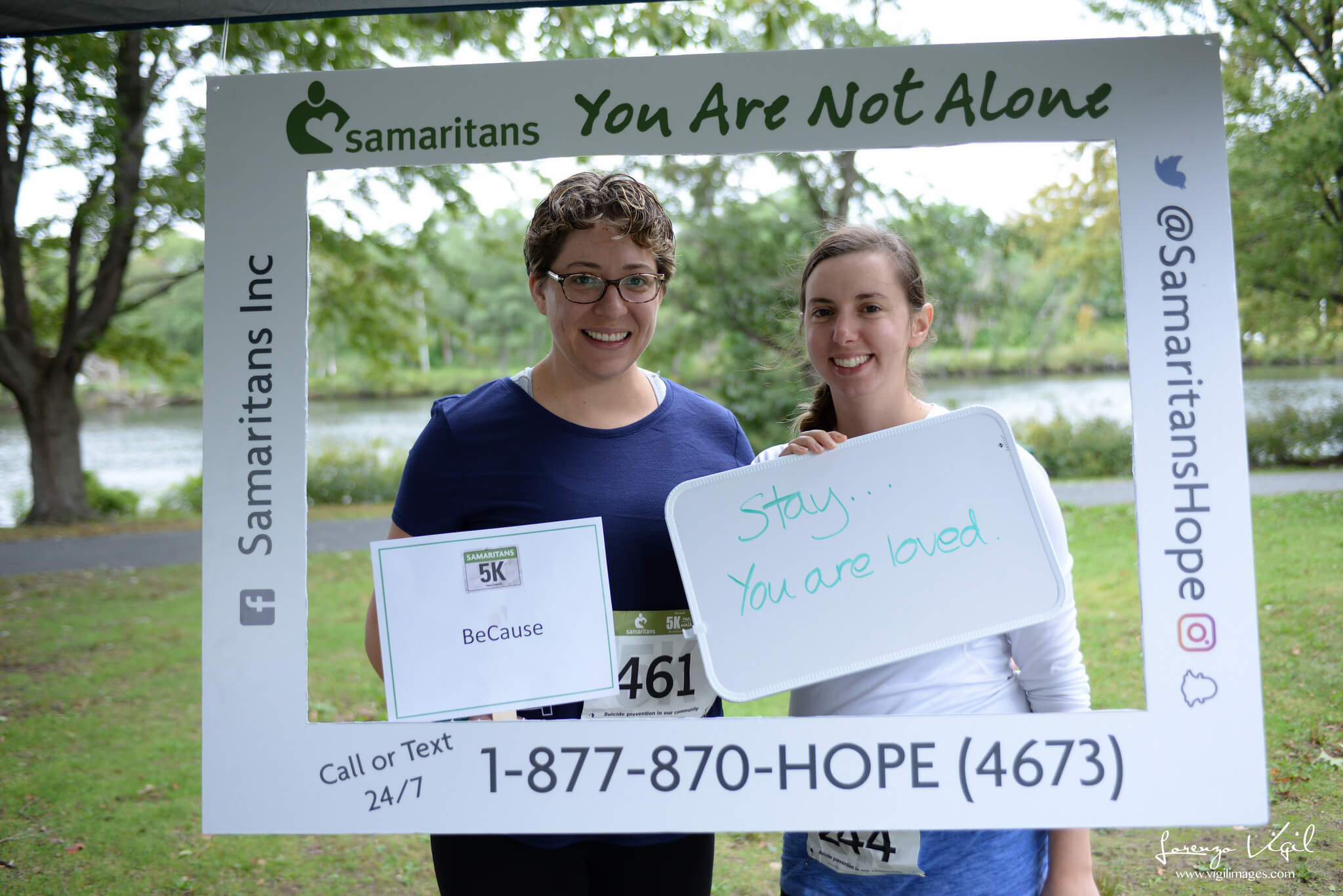 Two people stand behind a white frame with Samaritans contact information at the 2017 annual 5K Run/Walk for Suicide Prevention; they hold their team name, BeCause, and a white board which reads "Stay... you are loved."