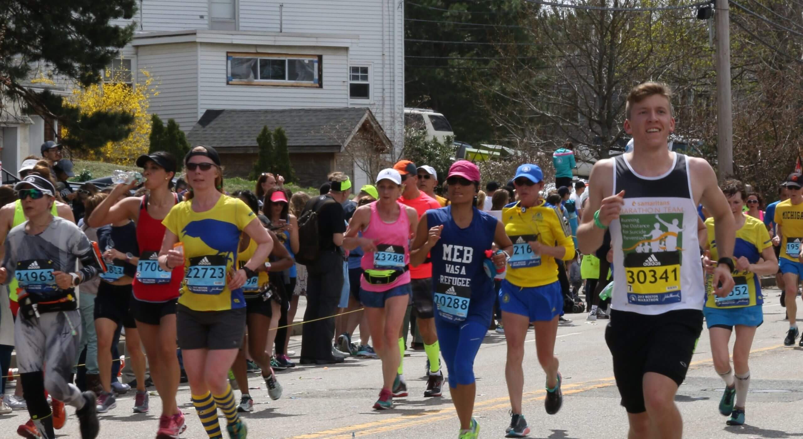 A group of Boston Marathon runners in the middle of the race. Most are wearing shorts, T-shirts and running bibs. Spectators and a white clapboard building are in the background.