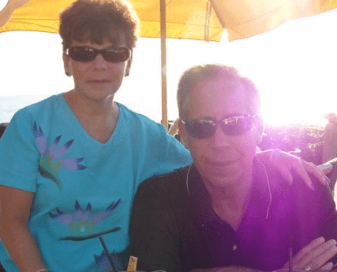 two people wearing sunglasses outside at a table under an umbrella