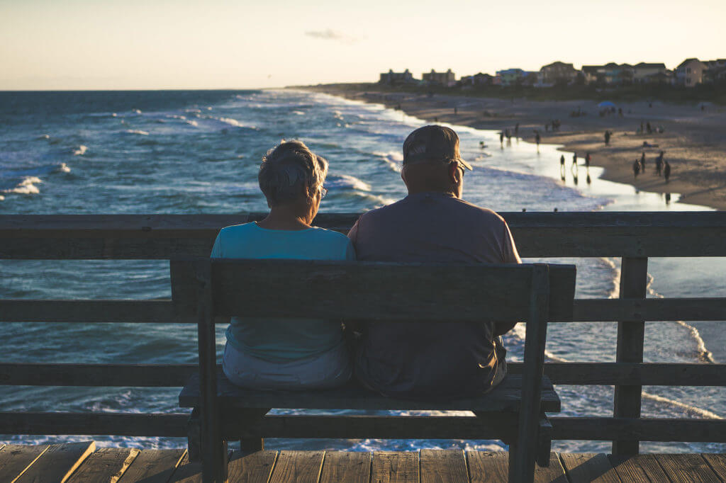 An older couple sitting on a bench on a pier by the beach.