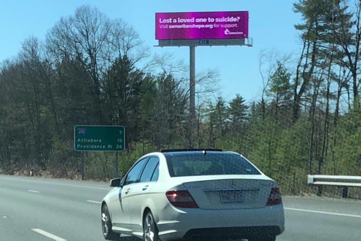 a white car on the highway passes a purple billboard with Samaritans information